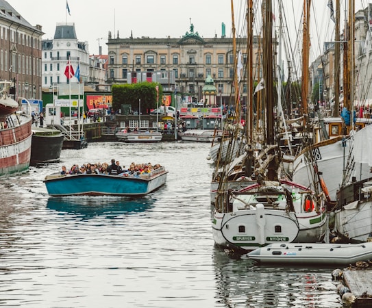 A lively harbor scene with numerous boats docked along a canal. In the foreground, a blue tour boat filled with people is cruising down the water. On either side of the canal, there are old brick buildings and vibrant storefronts, including visible flags. The atmosphere suggests a bustling and popular tourist area.