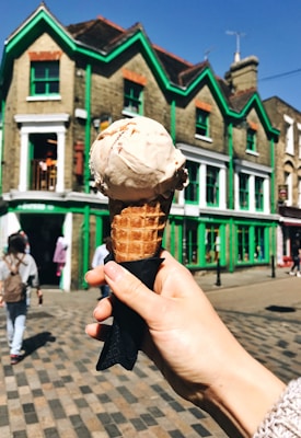A hand holding an ice cream cone with a single scoop is prominently displayed in the foreground. The background features a charming street scene with a distinctive building painted with green trim. People are casually walking along the cobblestone street under a clear blue sky.