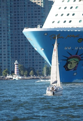 Carnival Cruises from Norfolk: 2026 & 2027 Sunshine, Magic, Freedom 3 A large cruise ship with vibrant artwork on its bow, featuring a stylized version of the Statue of Liberty, is juxtaposed with a small sailing boat in the foreground. The background is dominated by tall, densely packed urban buildings, highlighting a mix of urban life and maritime travel.