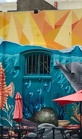 The Weird Norfolk Walk: 90-Minute Loop From the Norfolk Cruise Terminal 2 A vibrant mural depicting an underwater scene with marine life, including a seahorse, fish, and dolphins. The mural also features colorful abstract patterns, bubbles, and coral. In front of the mural, there are tables with red umbrellas and some parked vehicles.