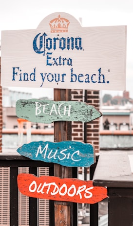 The Weird Norfolk Walk: 90-Minute Loop From the Norfolk Cruise Terminal 5 A wooden signpost stands by a waterfront, featuring a promotional message for Corona Extra with the phrase 'Find your beach.' Underneath, colorful wooden signs labeled 'BEACH,' 'MUSIC,' and 'OUTDOORS' are attached. The backdrop includes a brick pillar and an industrial or urban waterfront area with buildings and boats.