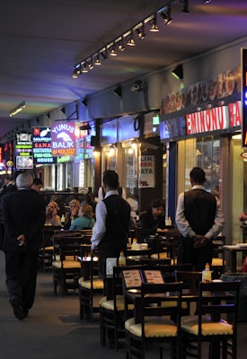 The Weird Norfolk Walk: 90-Minute Loop From the Norfolk Cruise Terminal 3 A bustling waterfront restaurant area underneath a bridge, with vibrant neon signs advertising food such as fish sandwiches. Several people are seated at outdoor tables enjoying their meals, while waitstaff in uniforms attend to them. The setting appears lively with a mix of locals and tourists.